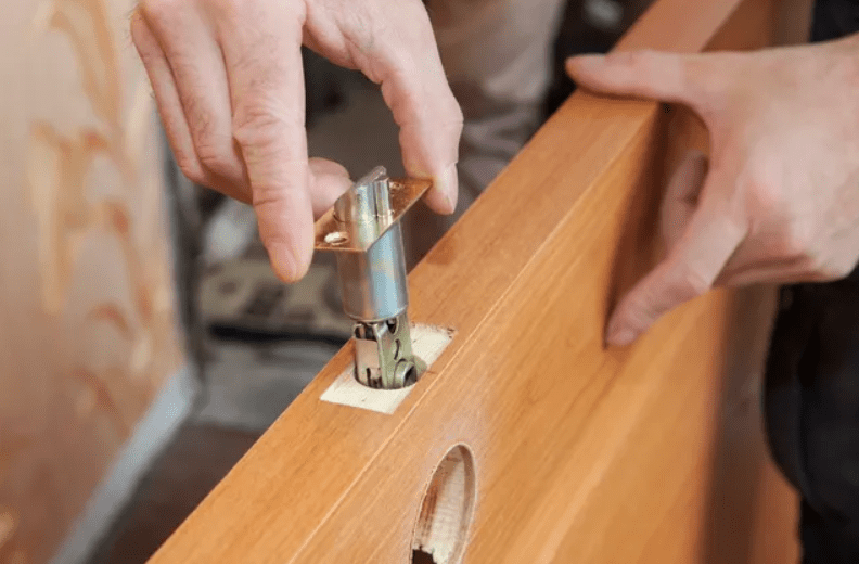 A person is installing a lock mechanism into a wooden door, inserting the latch into a cutout on the door's edge.