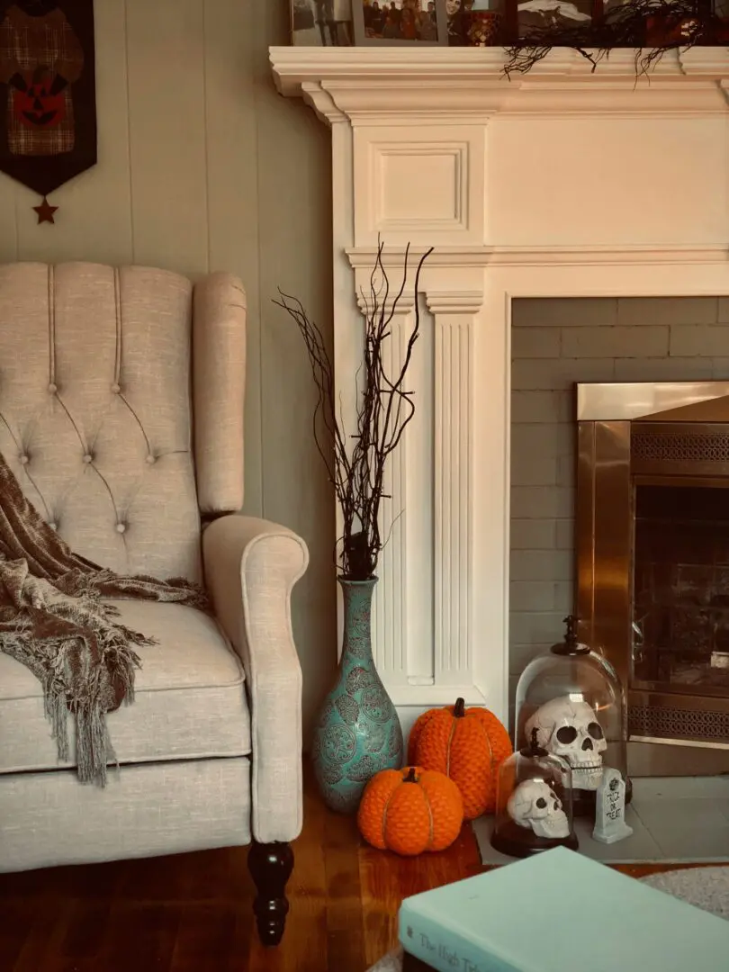 A beige armchair with a gray throw sits next to a fireplace. Two pumpkins, a turquoise vase with twigs, and a dome with skulls are on the floor beside it. A book is on a wooden floor.