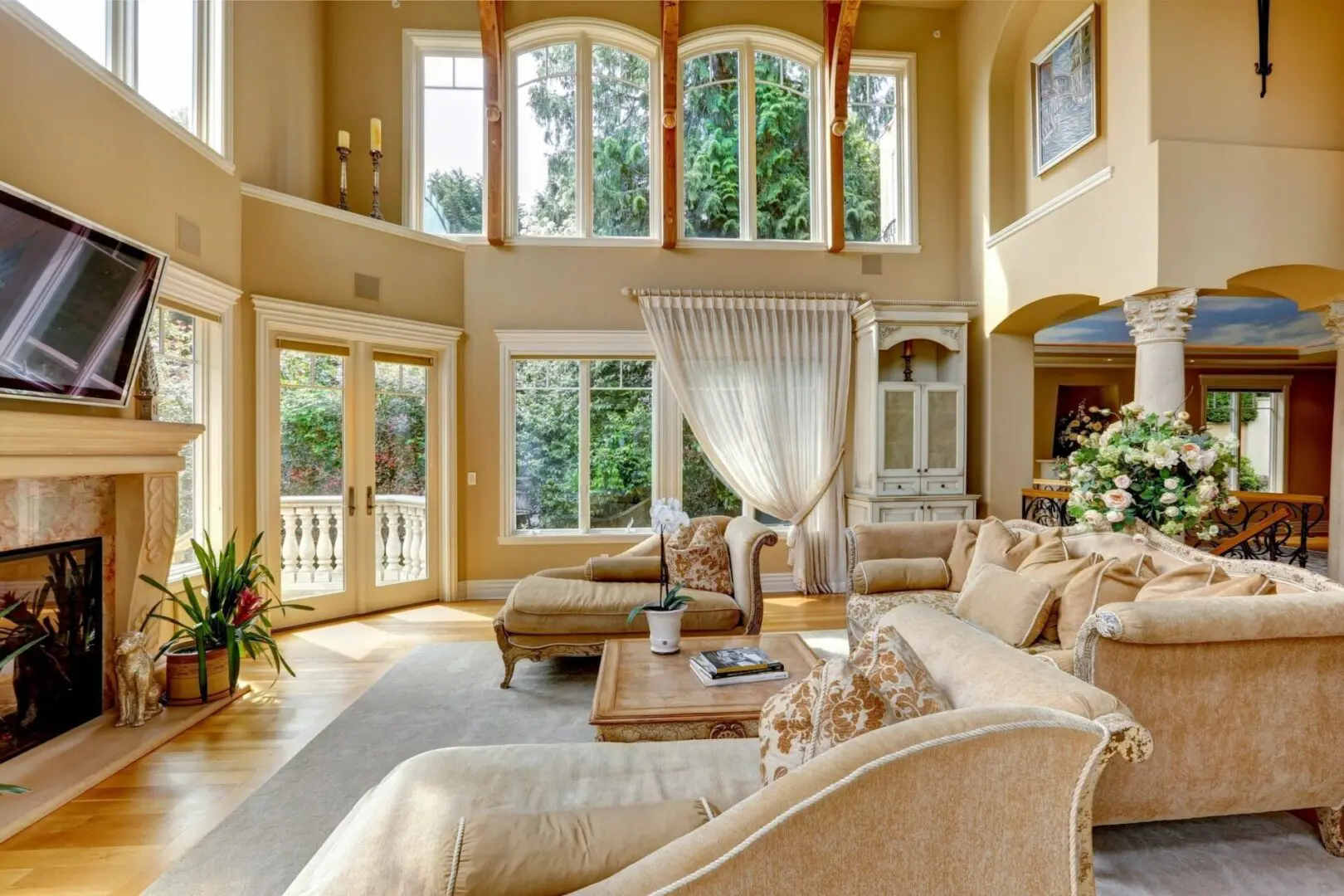 Spacious living room with high ceilings, large arched windows, beige sofas, a coffee table, a mounted TV above a fireplace, and a view of greenery outside.