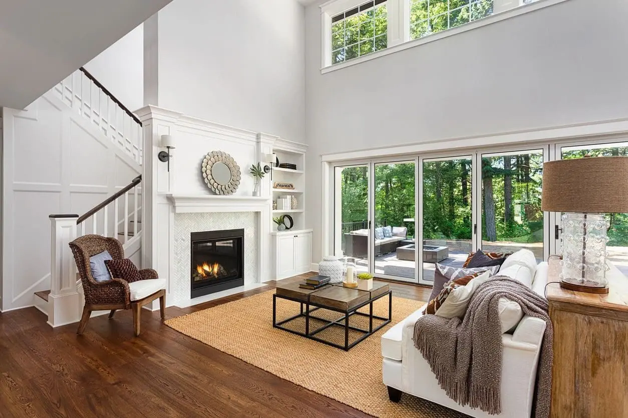 A modern living room with a fireplace, large windows, a white couch, a wooden armchair, and a coffee table on a rug. There is greenery visible outside the sliding glass doors.