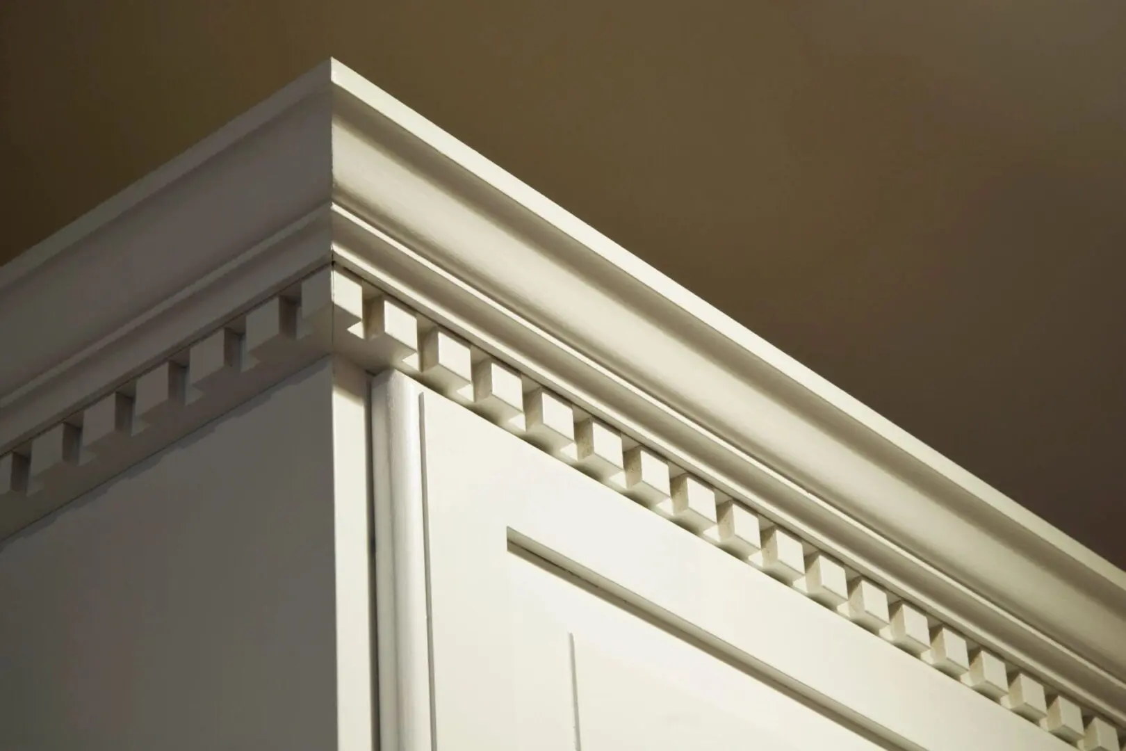 Close-up of white crown molding with detailed dentil trim at the top of a cabinet. The background shows a beige wall and ceiling.