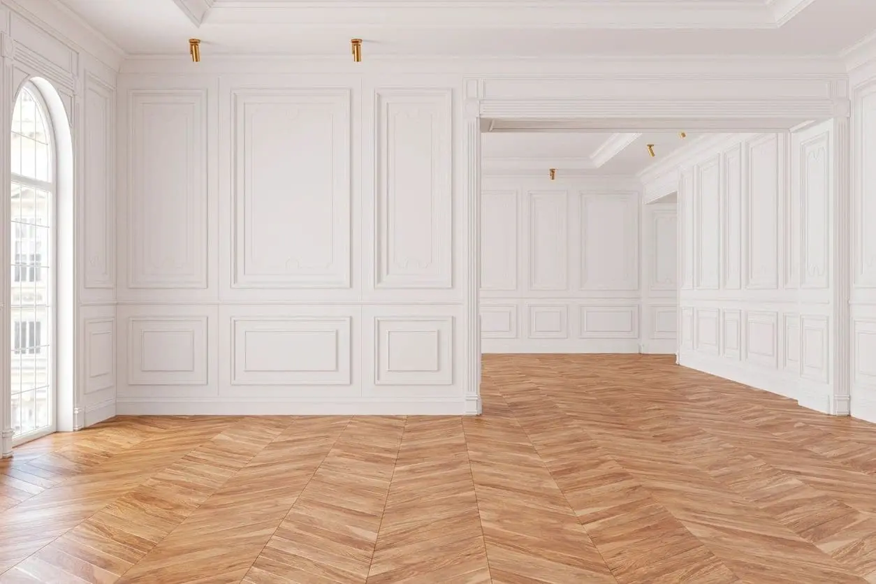 Spacious empty room with white paneled walls, wooden flooring in a herringbone pattern, and arched windows letting in natural light.