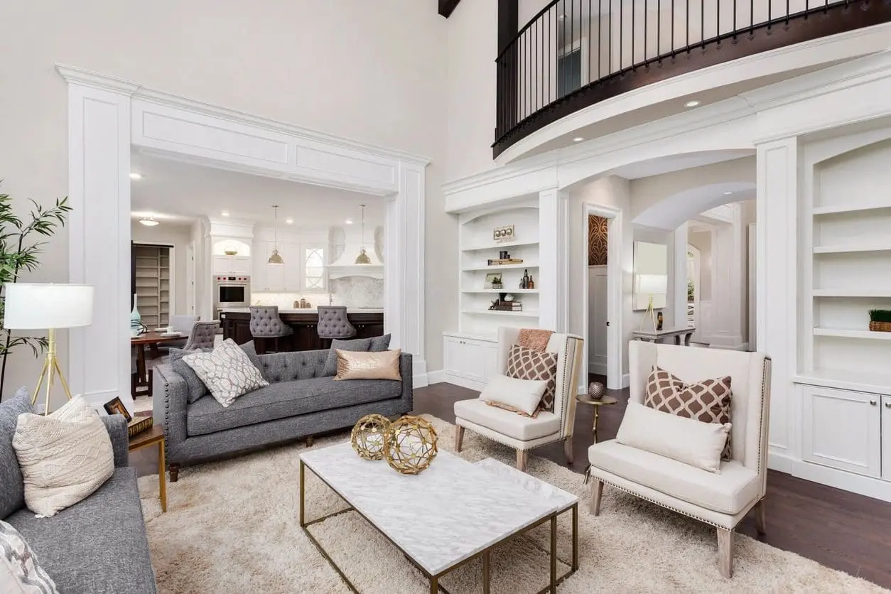 Spacious modern living room with gray sofas, white armchairs, a marble coffee table, and built-in shelves. An upper loft overlooks the open layout, leading to a well-equipped kitchen area.