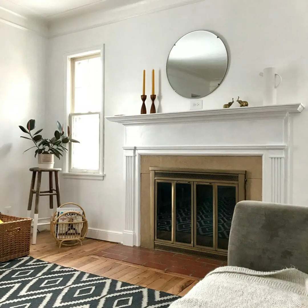 A living room with a white mantel above a fireplace, a round mirror, two candles, a small plant on a stool, a basket on the floor, and a patterned rug.