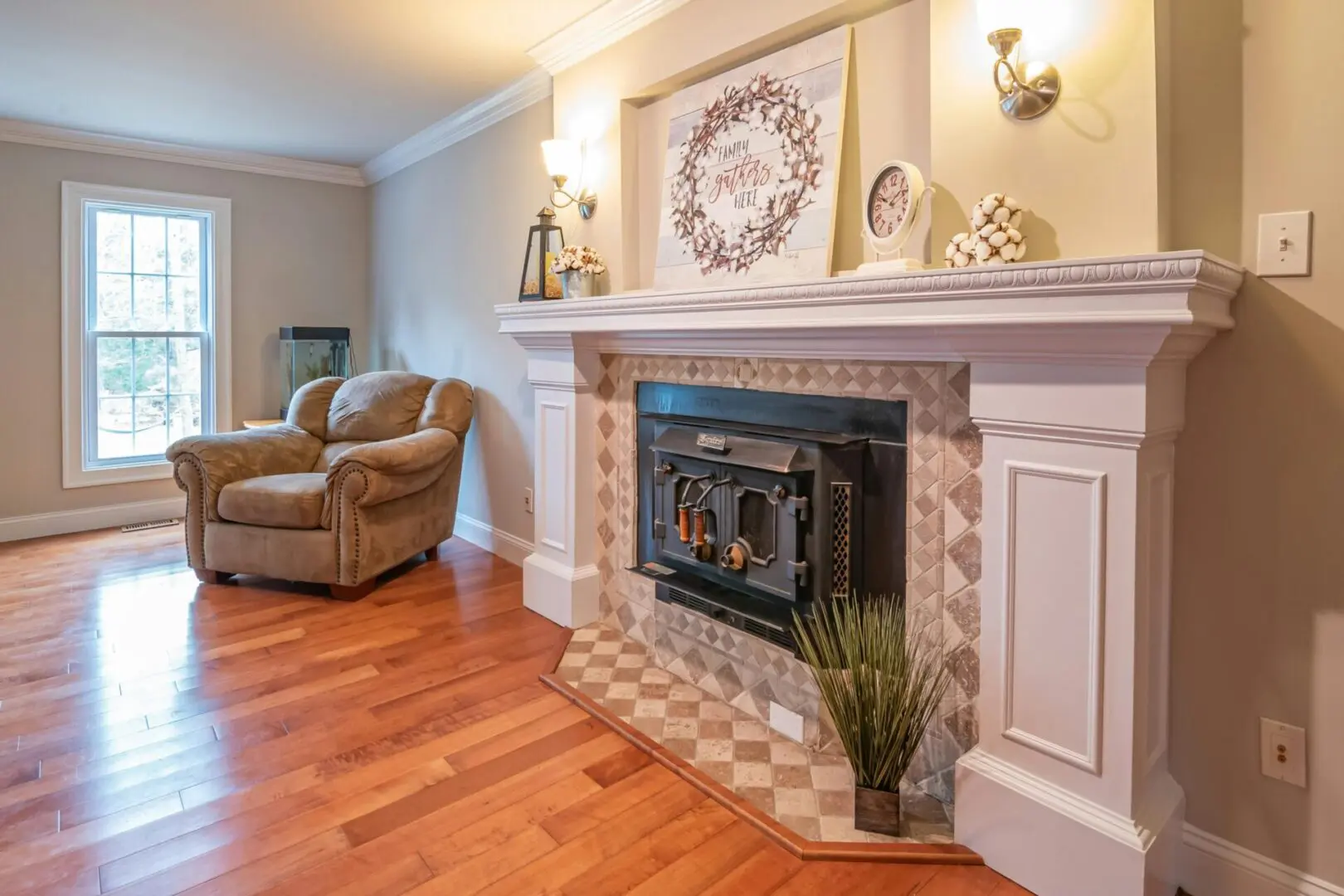 A cozy living room features a plush armchair next to a large window and a white fireplace decorated with a wreath, framed artwork, and a clock. The floor is hardwood, and the walls are a soft beige.