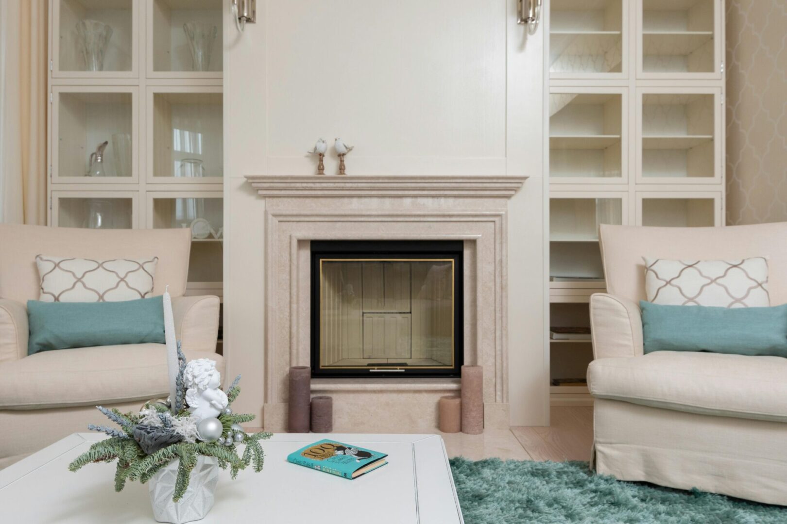 A living room with a beige fireplace, two beige armchairs with blue cushions, a coffee table with a decorative centerpiece and a book, built-in shelves, and a blue rug.